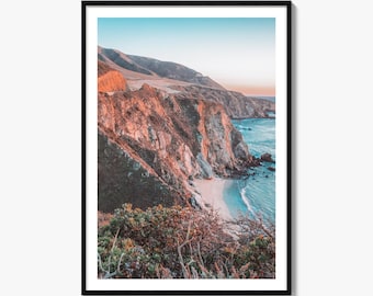 May include: A framed landscape photograph featuring a rugged coastal cliffside illuminated by warm golden hour light, overlooking a turquoise ocean and a small sandy beach, with dark green coastal shrubs in the foreground under a clear sky.