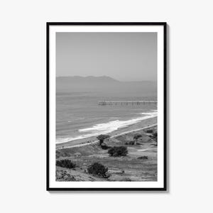 May include: A black and white framed wall art print featuring a coastal landscape. The scene displays a long pier extending into the ocean, a sandy beach with gentle waves, and rolling hills covered in sparse vegetation and scattered trees.