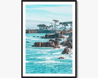 May include: A framed vertical wall art print featuring a coastal landscape with turquoise ocean waves crashing against jagged brown rocks. In the background, a line of cypress trees stands along a cliff under a bright, pale blue sky.