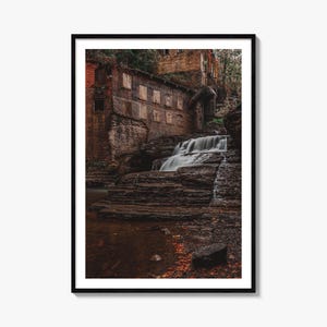 May include: A framed wall art print featuring a weathered brick building with boarded-up windows situated beside a cascading waterfall. The scene includes dark layered rock formations, flowing white water, and a pool with scattered autumn leaves.