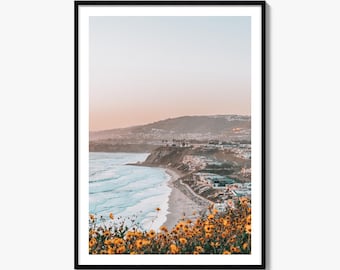 May include: A framed vertical wall art print featuring a coastal landscape at sunset. The foreground is filled with vibrant yellow wildflowers, overlooking a sandy beach, turquoise ocean waves, and a cliffside town under a soft pastel sky.