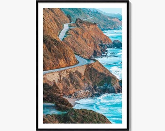 May include: A framed vertical wall art print featuring a winding coastal highway carved into steep, golden-brown cliffs. The road is bordered by a stone retaining wall, overlooking a bright turquoise ocean with white waves crashing against rocks.