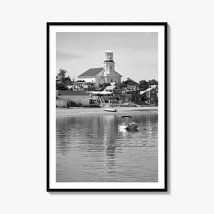 May include: A black and white framed wall art print featuring a coastal scene with a small motorboat floating on calm water in the foreground. In the background, a white church with a tall steeple stands above a cluster of houses near the shore.