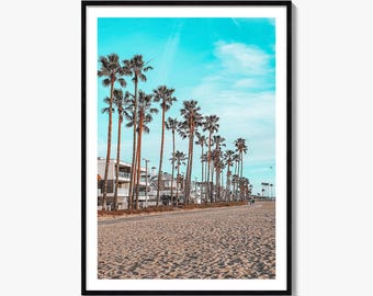 May include: A framed wall art print featuring a sandy beach with a row of tall palm trees stretching into the distance under a bright cyan sky. Multi-story residential buildings are visible along the shoreline next to the sandy path.
