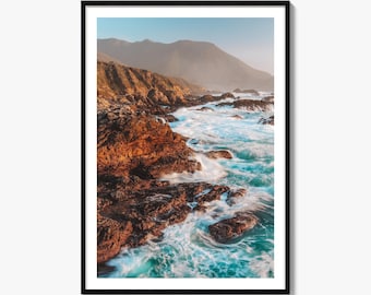 May include: A framed vertical wall art print featuring a coastal landscape with rugged, dark brown rocky cliffs meeting turquoise ocean waves. In the background, soft, hazy mountains rise under a clear, pale blue sky during the golden hour.