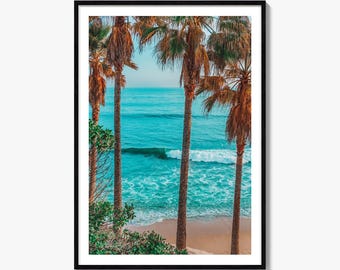 May include: A framed vertical wall art print featuring a view of a turquoise ocean with gentle waves rolling onto a sandy beach, framed by tall, slender palm trees with brown trunks and green fronds under a clear, light blue sky.