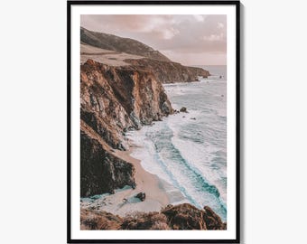 May include: A framed wall art print featuring a coastal landscape with rugged brown cliffs meeting a sandy beach and the ocean. The scene captures waves crashing against the shore under a soft, hazy sky with muted pink and golden light tones.