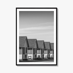 May include: A black and white framed wall art print featuring a row of identical small houses with gabled roofs and shuttered windows, arranged in a linear perspective against a clear sky, captured in a high-contrast monochromatic photographic style.