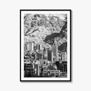 May include: A black and white framed wall art print featuring a dense urban cityscape nestled at the base of a massive, rugged mountain range. The composition highlights a prominent domed skyscraper surrounded by various mid-rise buildings.