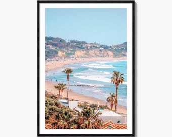 May include: A framed vertical wall art print featuring a coastal landscape with a sandy beach, turquoise ocean waves, and palm trees in the foreground. The background shows cliffs with residential buildings under a clear, bright blue sky.