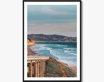 May include: A framed vertical wall art print featuring a coastal landscape with a concrete bridge extending over a sandy beach and ocean waves. The scene includes a distant hillside with buildings under a soft blue and orange sunset sky.