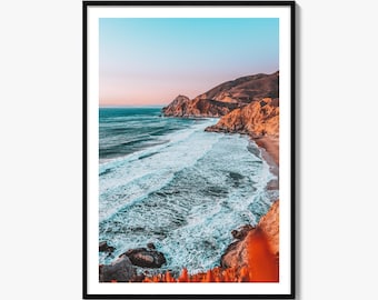 May include: A framed vertical wall art print depicting a coastal landscape at sunset. The scene features turquoise ocean waves crashing against a rocky shoreline with golden-brown cliffs under a gradient sky transitioning from soft pink to light blue.
