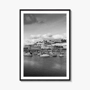 May include: A black and white framed wall art print depicting a harbor scene with numerous sailboats moored in calm water. In the background, a coastal town features multi-story buildings and a prominent church spire under a cloudy sky.