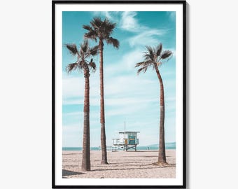 May include: A framed wall art print featuring three tall palm trees standing on a sandy beach under a bright blue sky with wispy clouds. In the background, a white wooden lifeguard tower sits near the shoreline, captured in a clear, daylight scene.