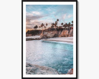 May include: A framed wall art print featuring a coastal landscape with a sandy beach, turquoise ocean water, and rocky cliffs topped with palm trees under a soft, pastel-colored sky with wispy clouds during the golden hour of sunset.