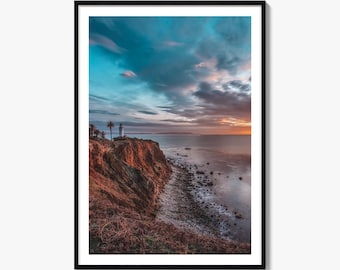 May include: A framed landscape photograph featuring a white lighthouse perched on a rugged cliffside overlooking the ocean at sunset. The sky displays a gradient of deep blue clouds transitioning into a warm orange glow above the horizon line.