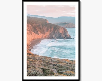 May include: A framed vertical wall art print featuring a coastal landscape with rugged brown cliffs meeting a turquoise ocean. The foreground shows textured green and yellow vegetation, while the background displays soft, hazy mountains under a pale pink sky.
