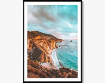 May include: A framed vertical wall art print featuring a coastal landscape with rugged orange-brown cliffs meeting a turquoise ocean. A bridge spans the cliffside above a sandy beach, all under a cloudy sky with soft light at the horizon.