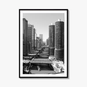 Può includere: Una stampa artistica da parete incorniciata in bianco e nero che raffigura un paesaggio urbano con alti grattacieli, tra cui due prominenti torri cilindriche, lungo un fiume. Diversi ponti attraversano l'acqua e una piccola barca è visibile.