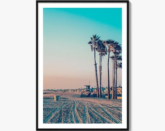 May include: A framed vertical wall art print depicting a sandy beach scene at sunset. The image features tall palm trees on the right, a lifeguard tower in the distance, and tire tracks in the sand. The sky transitions from a soft peach to blue.