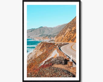 May include: A framed vertical wall art print depicting a winding coastal highway carved into a steep, orange-toned cliffside. The scene features a bright blue sky, deep blue ocean waves crashing against the rocks, and a wooden guardrail.