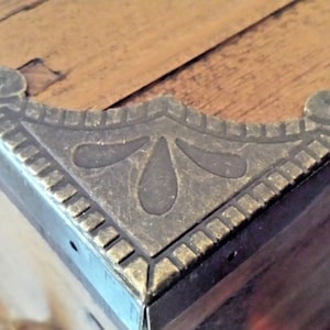 May include: A close-up of a brass corner brace with a decorative design. The brace is attached to a wooden surface.