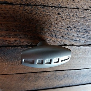 May include: A silver-coloured, oblong-shaped bottle stopper with a small handle. The stopper has three rectangular cutouts on one side. The object is resting on a wooden surface.