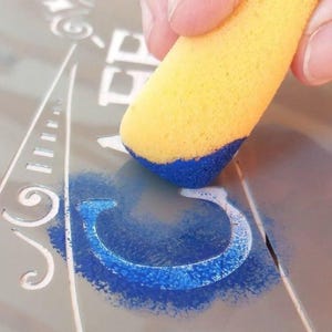 May include: A yellow sponge dauber is being used to apply blue paint to a stencil on a silver surface. The stencil is of the letter "C".