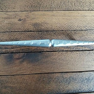 May include: A silver-coloured, double-ended tool with a series of graduated circular holes along each tapered end. The tool has a central, wider section with embossed circular designs. The tool is lying on a wooden surface.