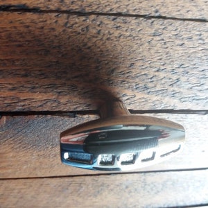 May include: A chrome cabinet knob with a wooden stem. The knob has a rectangular base with three cut-out sections. The wood stem is light brown. The knob is set against a dark brown wooden surface.