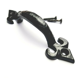 May include: Black metal door handle with a decorative fleur-de-lis design. The handle is attached to a white surface.