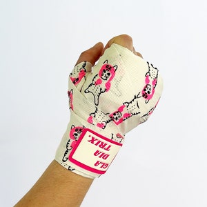 Puede incluir: Vendas de boxeo blancas con un estampado rosa y negro de gatos de dibujos animados. Las vendas tienen una etiqueta rosa que dice "GLA DIA TRIX".