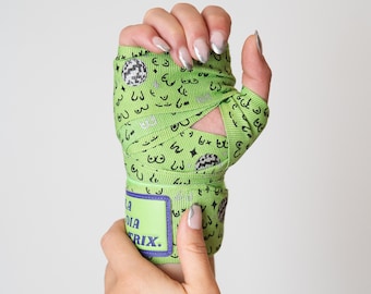 Puede incluir: Una venda verde para la mano con un patrón repetido de senos, bolas de discoteca y estrellas. La venda tiene un parche rectangular con el texto "LA DIATRIX" en blanco y morado. La venda está enrollada alrededor de una muñeca.