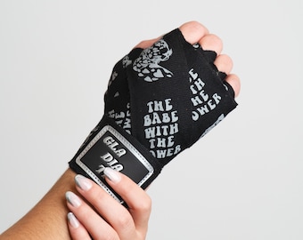 Puede incluir: Vendas de muñeca negras con un estampado plateado de un tigre y el texto "The Babe With The Power". Las vendas tienen una etiqueta plateada que dice "Gladia".