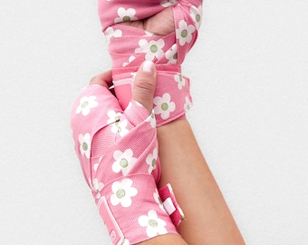 Puede incluir: Vendas de boxeo rosas con un estampado floral blanco. Las vendas están envueltas alrededor de las manos y las muñecas. El estampado floral blanco tiene centros verdes. Las vendas se aseguran con una correa de velcro rosa.