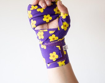 Puede incluir: Vendas de mano moradas con estampado floral amarillo. Las vendas están envueltas alrededor de un puño y una muñeca, con cierre de velcro. La tela presenta un diseño repetido de flores amarillas sobre un fondo morado. Diseñado para boxeo u otros deportes de combate.
