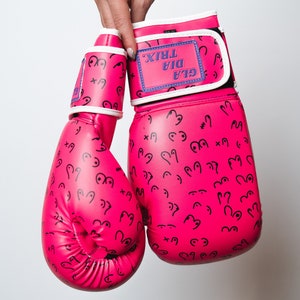 Puede incluir: Guantes de boxeo rosas con ribete blanco y un patrón de garabatos negros. Los guantes tienen una etiqueta blanca con el texto "GLA DIA TRIX" en morado.