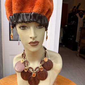 May include: An orange faux fur beret with a dark brown leather band is displayed on a mannequin. The mannequin also wears a brown wooden necklace with gold accents and matching earrings. The beret is a stylish accessory.