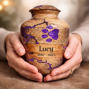May include: A wooden pet urn with a paw print design and the name "Lucy" and dates "2007-2023" engraved on it. The urn is brown with purple accents and is held in someone's hands.
