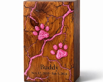 May include: A wooden pet urn with a pink paw print design and the name "Buddy" and dates "Nov 12, 2010 - Feb 5, 2024" engraved on the front. The urn is rectangular and made of dark wood with pink accents.