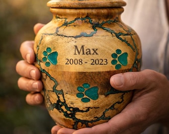 May include: A wooden pet urn with a lid, featuring a natural wood grain pattern with teal accents. The urn is engraved with the name "Max" and the years "2008-2023", along with teal paw prints. The urn is held in both hands.