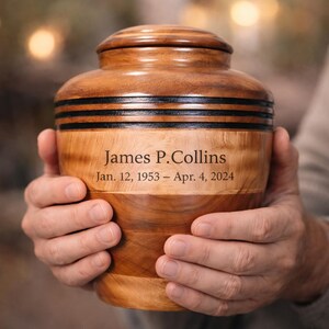May include: A wooden urn with a lid, held by two hands. The urn is a light brown color with dark brown bands. The text "James P. Collins" and dates "Jan. 12, 1953 &ndash; Apr. 4, 2024" are engraved on the front.