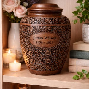 May include: A brown urn with intricate floral carvings and a lid. The urn has an oval plaque with the name "James Wilson" and the years "1958-2023". The urn is displayed on a wooden shelf with candles and flowers.