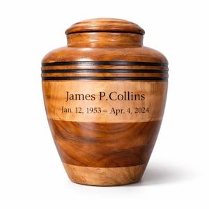 May include: A wooden urn with a lid, featuring a layered design of light and dark wood. The urn has a cylindrical shape with two black bands. The text "James P. Collins Jan. 12, 1953 &ndash; Apr. 4, 2024" is engraved on the front.