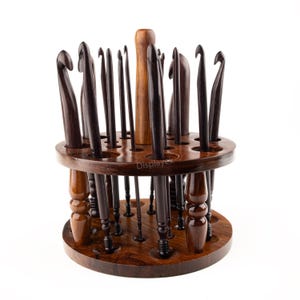 Peut inclure: Un porte-crochets en bois avec 24 fentes pour ranger les crochets à crochet. Le porte-crochets est en bois brun foncé et a une base ronde. Le porte-crochets est rempli de crochets à crochet de différentes tailles et couleurs.