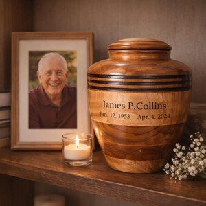 May include: A wooden urn with a lid, featuring dark bands and the engraved name "James P. Collins" and dates. A framed photo and a lit candle sit nearby on a wooden shelf. White flowers are to the right.