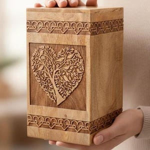 May include: A rectangular wooden cremation urn featuring a light brown natural wood grain finish. The front panel displays a carved heart-shaped tree design with intricate branches and leaves. Decorative knotwork borders line the top and bottom.