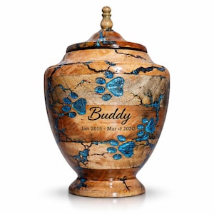 May include: A wooden pet urn with a lid, featuring a natural wood grain pattern and blue resin accents. The urn is adorned with blue paw prints and the name "Buddy" along with the dates Jan 2018 - Mar 5 2024.