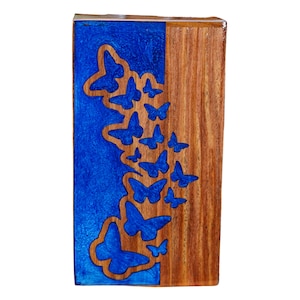 May include: A rectangular wooden box with a blue resin inlay featuring butterfly cutouts. The right side is natural wood grain. The butterflies are various sizes and shapes, creating a whimsical design. The box is likely for decorative or storage purposes.
