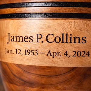 May include: A wooden urn with the name "James P. Collins" and the dates "Jan. 12, 1953 &ndash; Apr. 4, 2024" engraved on it. The urn is made of light and dark brown wood with black accents.